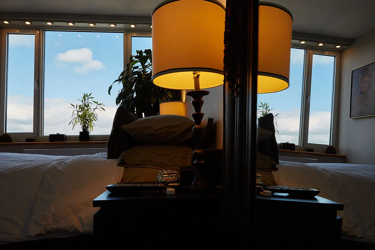 Bedroom with lit table lamp, plants and wide windows under blue sky, reflected in mirror